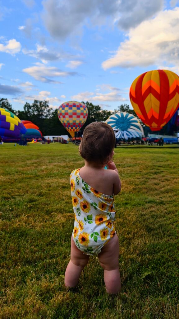 Toddler in sunflower onsie looking at hot air balloons inflating.
